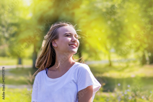 young cute beautiful girl 16 years old with long hair, smiling cheerfully, raising her face to the sun, female portrait of a teenager girl in nature in sunny weather