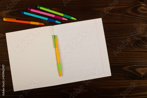 School notebook with oblique lines, multicolored pens on the table brown wooden background. Blank sheet of paper in notebook on the desk. Back to school. School supplies. Top view. Copy space for text