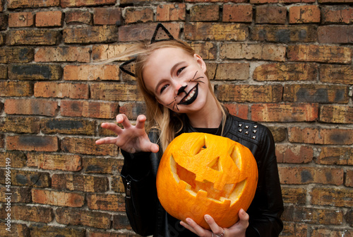Beautiful teenager girl dressed as a black cat for Halloween. Child holding a pumpkin monster for Halloween. 