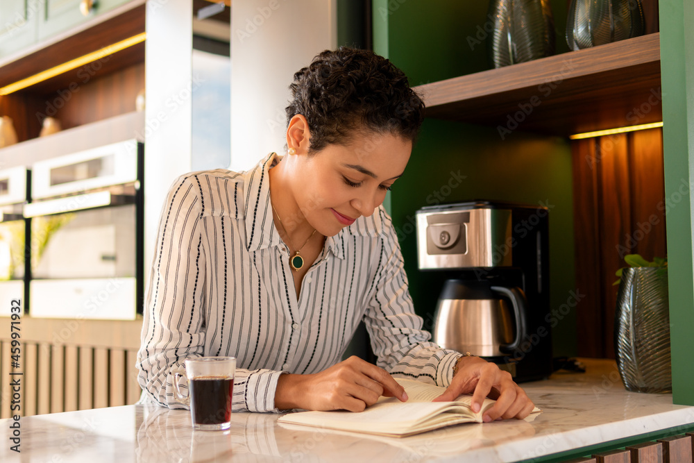 happy woman reading book and drinking coffee in kitchen counter