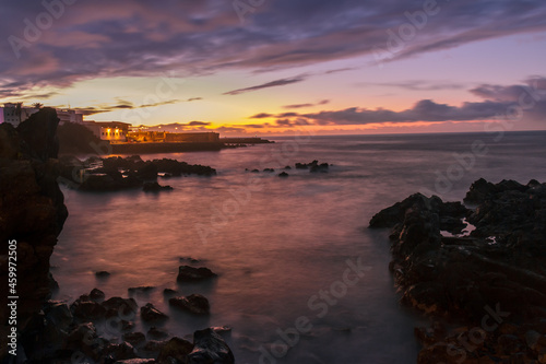 long exposure photograph of sea water flowing between the rocks into the ocean and some buildings in the background with the light of the sunset in the canary islands