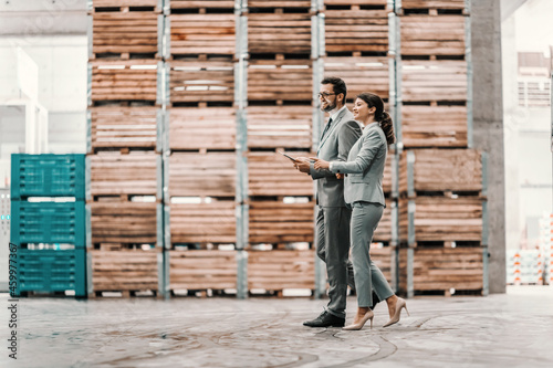 Business talk and walk in the warehouse. Man and a woman in a business suit stand in a warehouse and check the situation. Man holds a tablet in his hand and points a finger at the woman at something