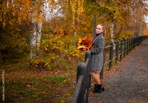 A blonde girl walks in the park in autumn. She is holding yellow leaves in her hands. She's happy. The sun is shining. Yellow leaves are lying nearby.