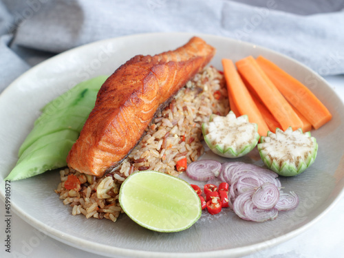 Close up of grilled salmon top on fried brown wild rice with spicy, Thai recipe. Side dish with mixed vegetables such as cucumber, carrots, Thai eggplant, shallot and lemon with red chili
