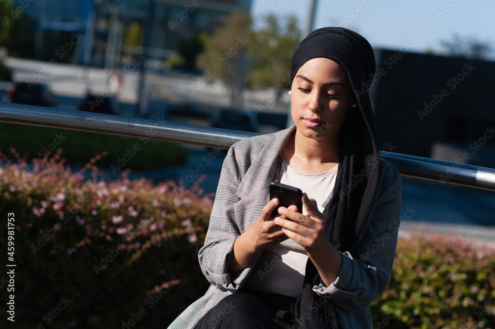 Adult woman wearing a black headscarf sitting on a modern park bench looking down using her phone Adult woman wearing a black headscarf sitting on a modern park bench looking down using her phone