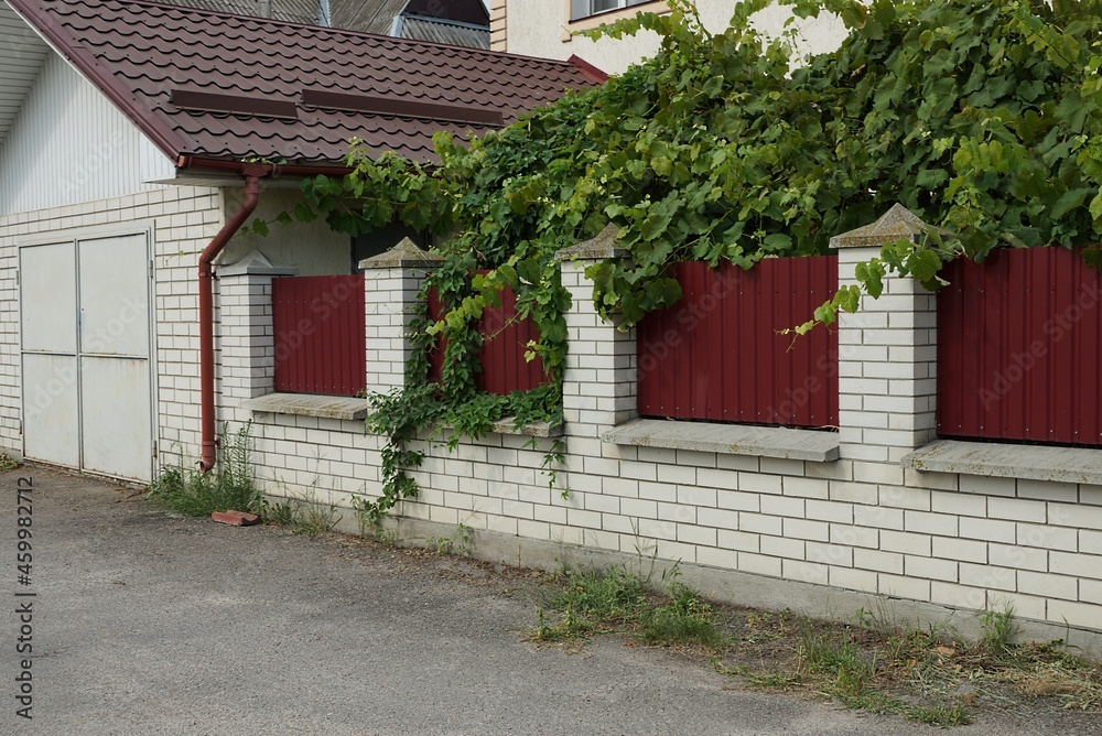 Naklejka premium garage facade with gray iron gates against the wall of a fence made of white bricks and red metal on the street in green vegetation