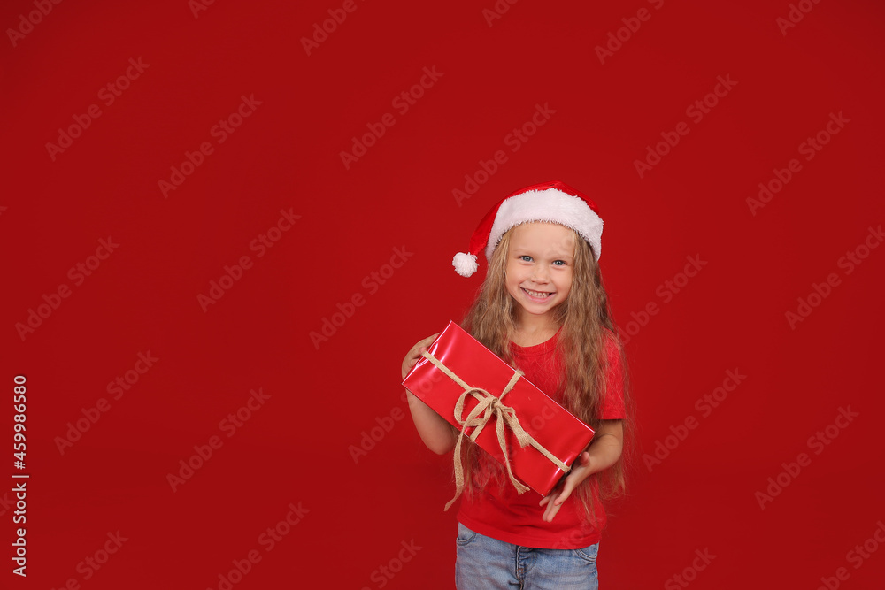 a beautiful happy little blue-eyed blonde girl in a red T-shirt with a Santa Claus hat on her head on a red background holds a new year gift box a beautiful happy little blue-eyed blonde girl in a red T-shirt with a Santa Claus hat on her head on a red background holds a new year gift box