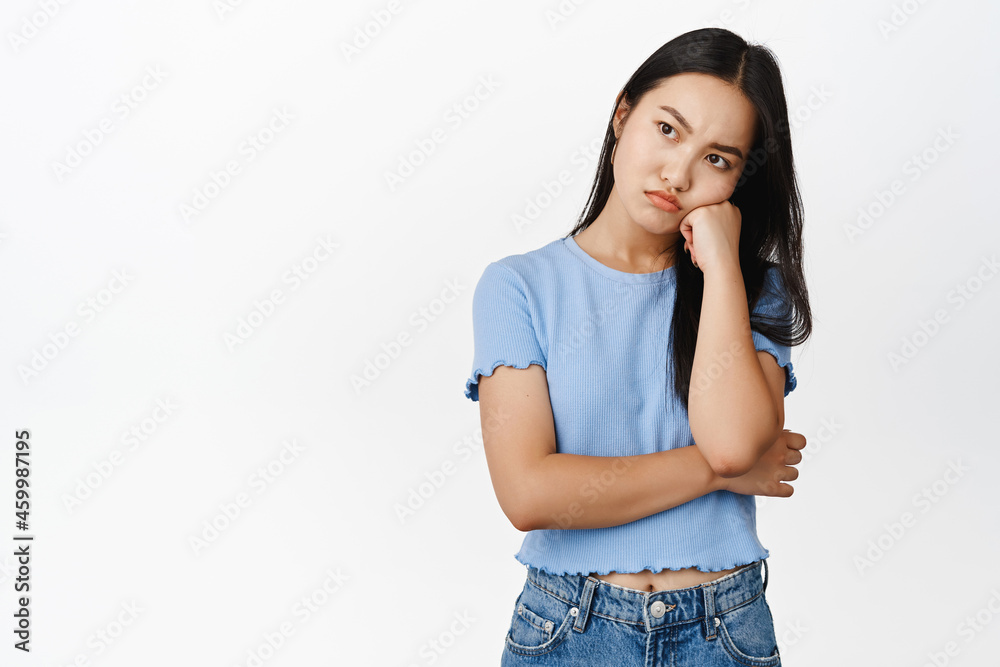 Angry asian girl lean on fist, looking aside with pissed-off moody face expression, standing insulated against white background