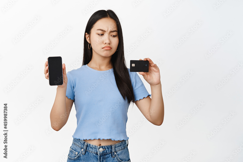 Disappointed asian girl showing mobile phone screen, looking upset at credit card, standing in blue t-shirt over white background Disappointed asian girl showing mobile phone screen, looking upset at credit card, standing in blue t-shirt over white background