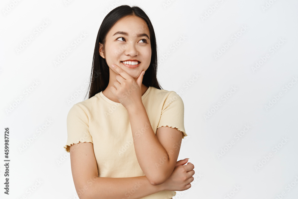 Obraz premium Smiling asian girl thinks, looks aside thoughtful, ponder something, standing over white background