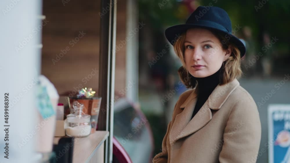 A beautiful lady in a brown coat and with a hat on her head stands on a sunny autumn day at the cafe bar and waits until the bartender makes coffee. Cheerful girl shows her tongue to camera and laughs A beautiful lady in a brown coat and with a hat on her head stands on a sunny autumn day at the cafe bar and waits until the bartender makes coffee. Cheerful girl shows her tongue to camera and laughs