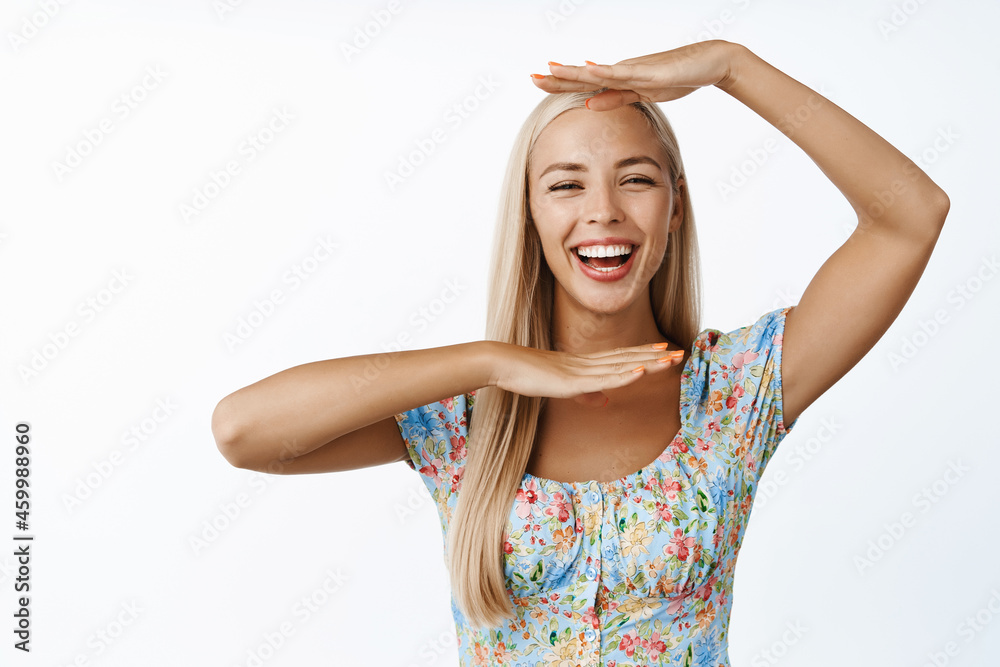 Happy blong girl laughs, shows her face, making hand frame and smiling, standing in dress over white background Happy blong girl laughs, shows her face, making hand frame and smiling, standing in dress over white background