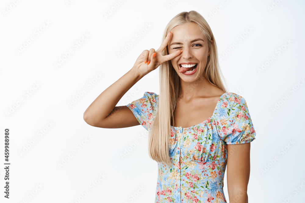 Beautiful blond girl winking, showing peace sign and tongue, coquettish pose, standing over white background Beautiful blond girl winking, showing peace sign and tongue, coquettish pose, standing over white background