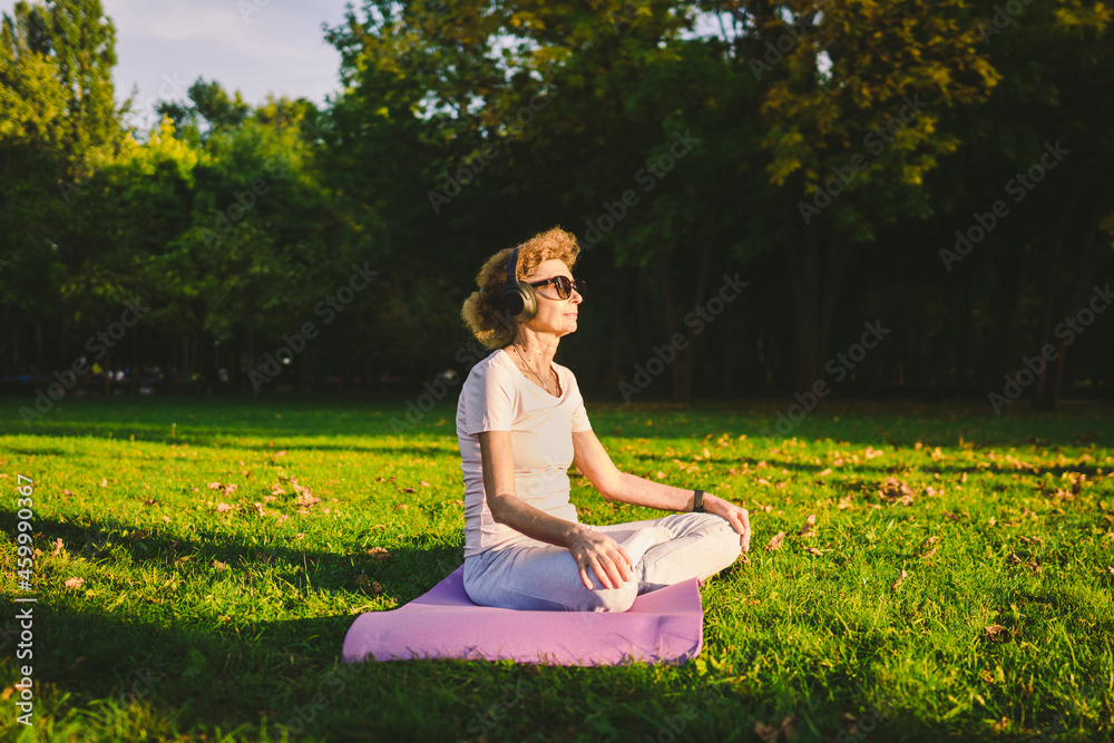 Senior woman meditating and exercising yoga lotus position outdoors. Ederly female doing stretching exercises on yoga mat in park on green lawn at sunset. Feeling energy. Concept calm and meditation Senior woman meditating and exercising yoga lotus position outdoors. Ederly female doing stretching exercises on yoga mat in park on green lawn at sunset. Feeling energy. Concept calm and meditation