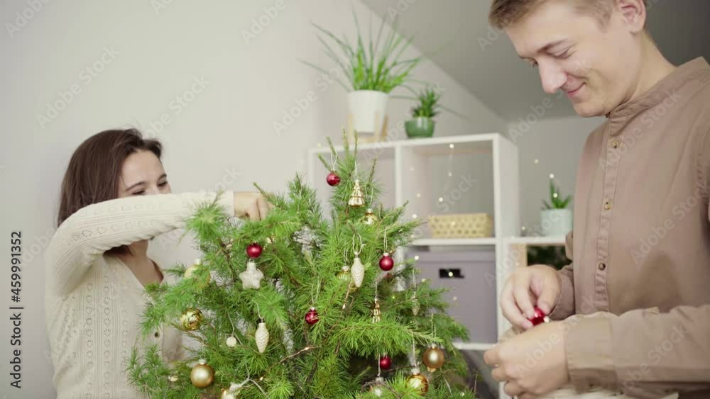 Happy family decorates the Christmas tree with red and gold balls in their home. A couple, a young woman and a man are preparing for the new year and Christmas holidays. Happy family decorates the Christmas tree with red and gold balls in their home. A couple, a young woman and a man are preparing for the new year and Christmas holidays.