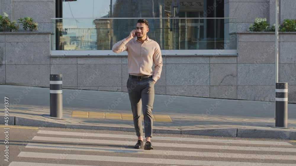 Hispanic arabic young adult business man walking with mobile phone at street pedestrian crossing road traffic. Boss businessman call smartphone outdoors. Cheerful guy talking cellphone outside going Hispanic arabic young adult business man walking with mobile phone at street pedestrian crossing road traffic. Boss businessman call smartphone outdoors. Cheerful guy talking cellphone outside going