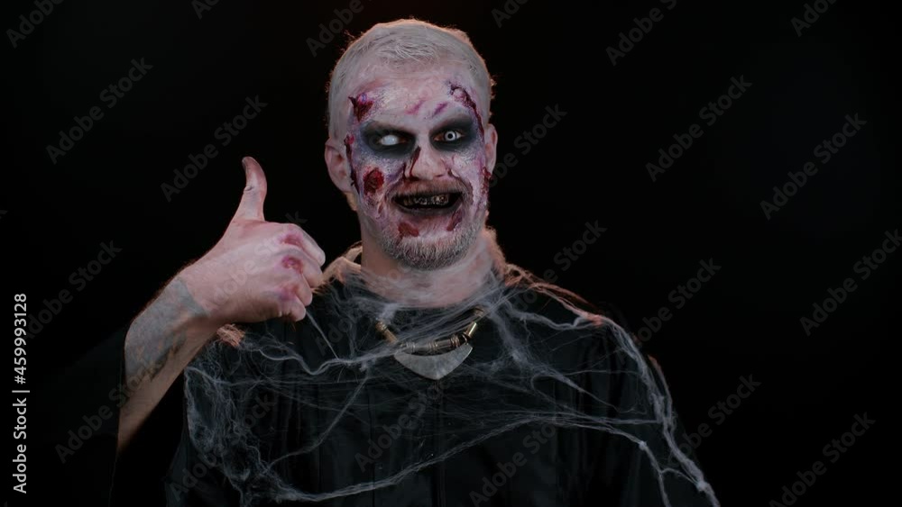 Sinister man with scary Halloween zombie makeup in costume raises thumbs up agrees with something or gives positive reply recommends advertisement likes good. Dead guy with wounded bloody scars face Sinister man with scary Halloween zombie makeup in costume raises thumbs up agrees with something or gives positive reply recommends advertisement likes good. Dead guy with wounded bloody scars face