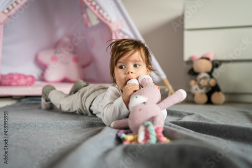 Baby small caucasian girl five months old lying belly down on tummy on the floor at home in day playing with toy in her mouth childhood concept alone front view leisure