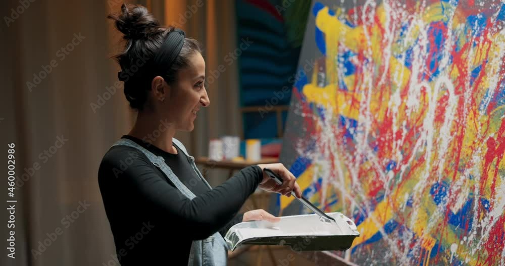 A woman with soul of artist is standing by canvas with abstract painting, holding container, paint tray and brush on long handle, mixing white paint and adding spots of it on canvas in specific places A woman with soul of artist is standing by canvas with abstract painting, holding container, paint tray and brush on long handle, mixing white paint and adding spots of it on canvas in specific places
