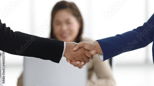 Close up shot of handshaking of unrecognizable unidentified successful businessman and businesswoman in formal suit in front female officer staff working with laptop computer in blurred background