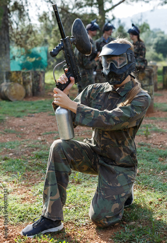Young woman in camouflage holding gun before paintball match with friends on open field