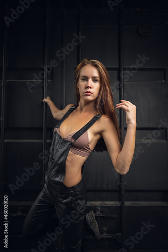 Outdoor close-up portrait of a young beautiful woman in a leather overalls and top posing against of an industrial  container.Fashion concept. © Виталий Сова