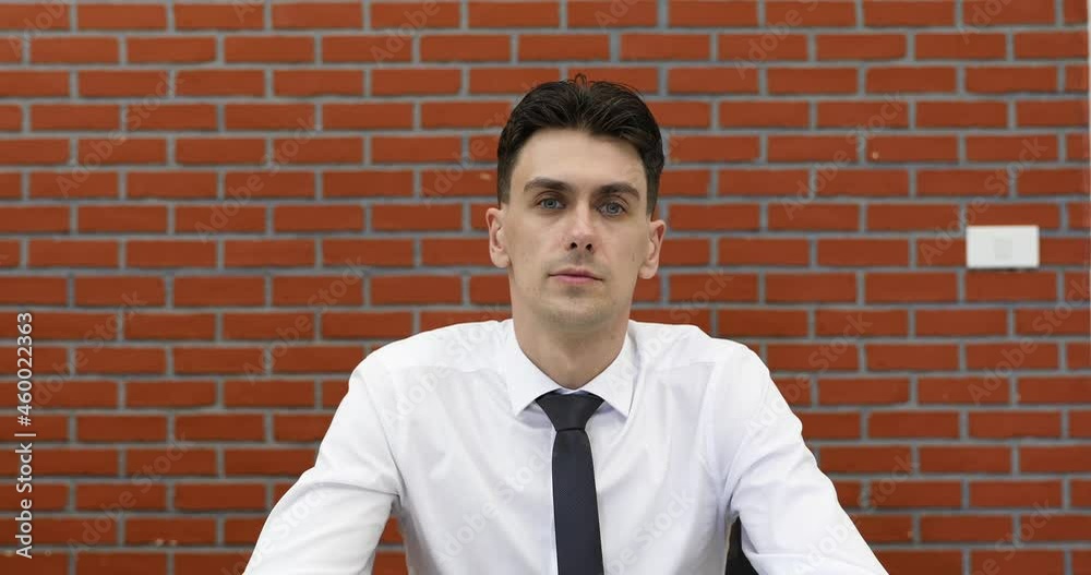 Young adult businessman in casual white shirt sitting in front of red brick wall listen to speaker in online conference and nod head with agreed and then raise hand for ask question. Young adult businessman in casual white shirt sitting in front of red brick wall listen to speaker in online conference and nod head with agreed and then raise hand for ask question.