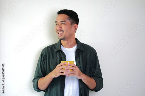 Adult Asian man looking side and smiling relax while holding a cup of morning coffee photo