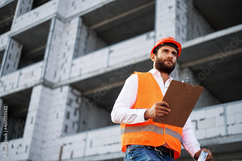 Young man engineer in workwear standing in construction site with clipboard