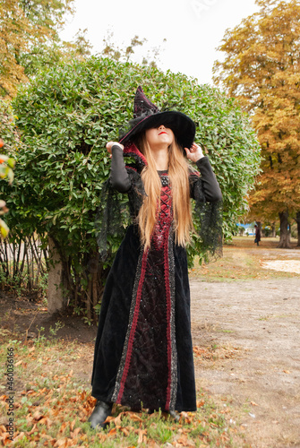 a girl in a witch costume for Halloween holding on to a hat with her hands hides her face against the background of greenery on Halloween