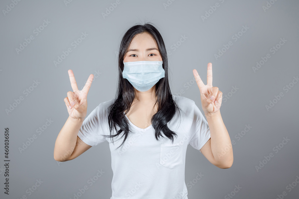 Portrait of Asian woman in white t-shirt, medical face mask to protect Covid-19 (Coronavirus) showing peace or victory gesture with both hands on gray background Portrait of Asian woman in white t-shirt, medical face mask to protect Covid-19 (Coronavirus) showing peace or victory gesture with both hands on gray background