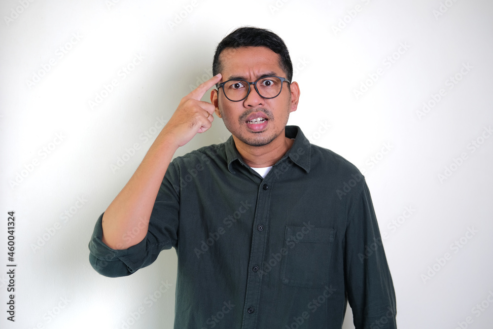 Adult Asian man showing angry expression with his finger pointing to side forehead Adult Asian man showing angry expression with his finger pointing to side forehead