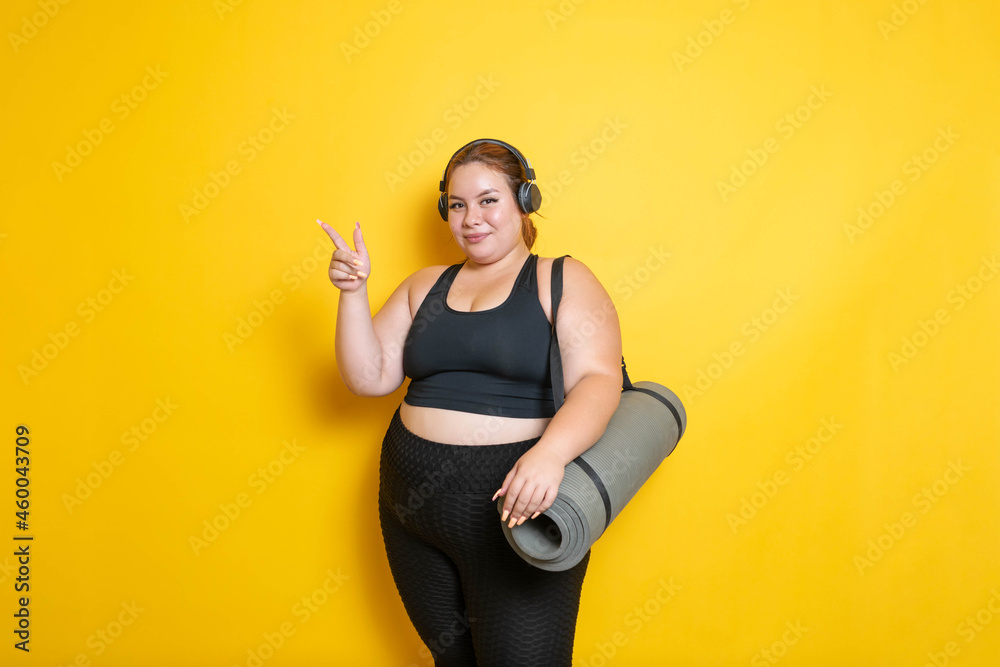 overweight and fitness young Caucasian Hispanic model in sportswear with headphones showing yoga mat pointing to the side overweight and fitness young Caucasian Hispanic model in sportswear with headphones showing yoga mat pointing to the side