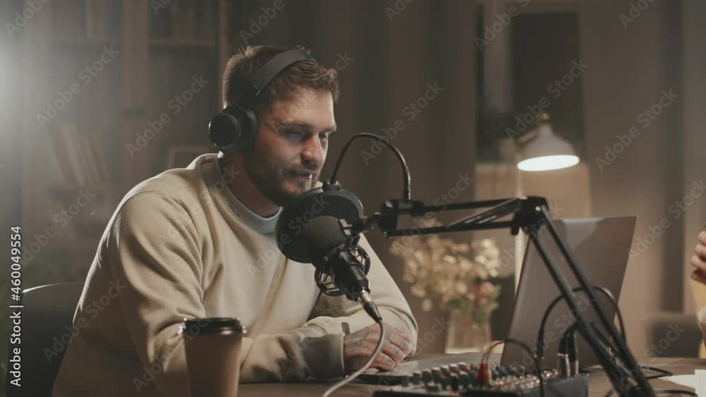 Handheld tracking of man in headphones sitting at table in living room lit by lamp and studio lights and talking into microphone while hosting online podcast together with unrecognizable woman Handheld tracking of man in headphones sitting at table in living room lit by lamp and studio lights and talking into microphone while hosting online podcast together with unrecognizable woman