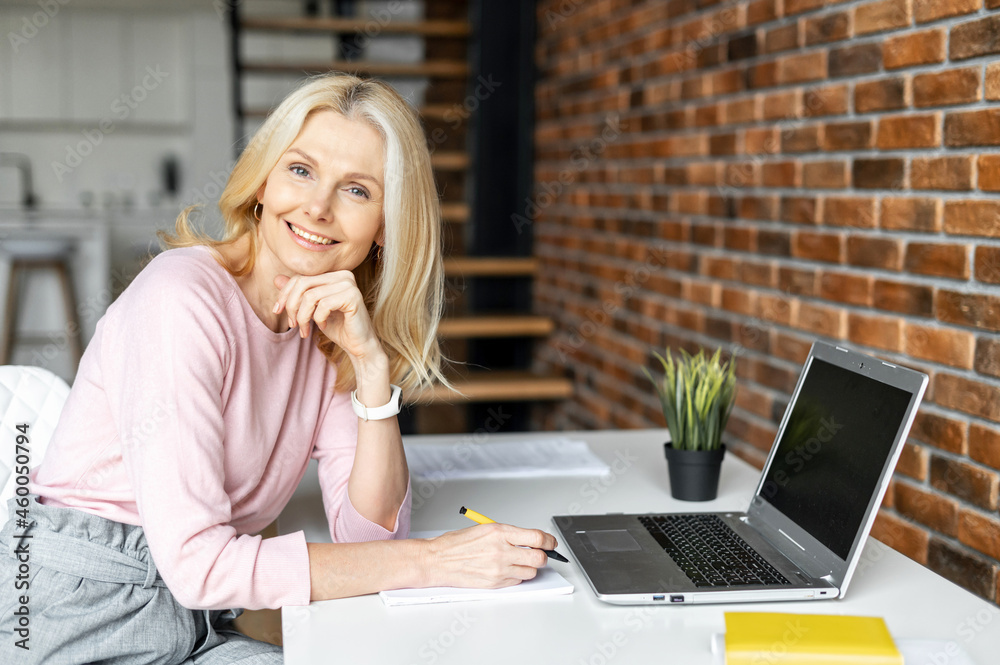 Positive charming mature blonde businesswoman sitting at the desk in front of laptop, working on a project. Freelancer enjoying remote work from home, looks at the camera and smiling Positive charming mature blonde businesswoman sitting at the desk in front of laptop, working on a project. Freelancer enjoying remote work from home, looks at the camera and smiling