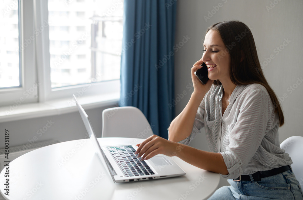 Work from home concept. Young female entrepreneur chatting on the smartphone sitting at the desk and using laptop, smiling woman has pleasant phone conversation during remote work Work from home concept. Young female entrepreneur chatting on the smartphone sitting at the desk and using laptop, smiling woman has pleasant phone conversation during remote work