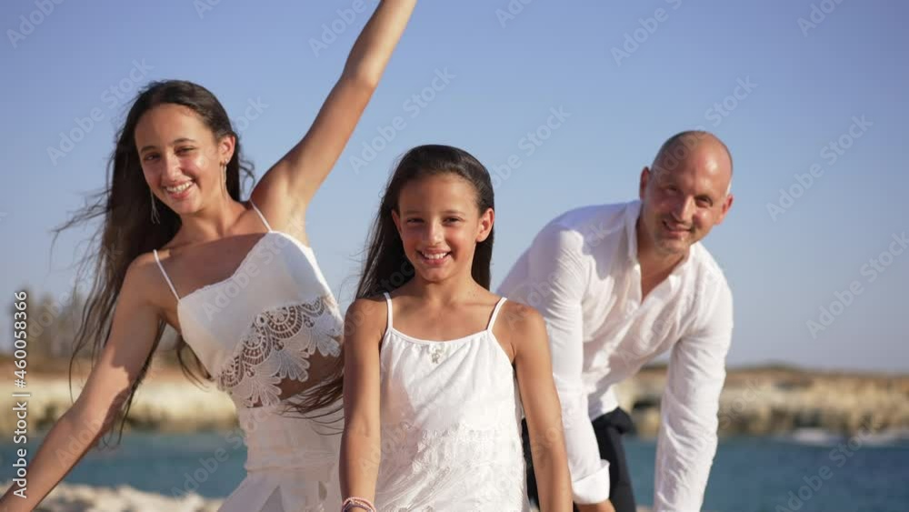 Joyful little girl smiling looking at camera as teenage sister and father bending to sides posing. Portrait of happy pretty Caucasian child standing with family on Mediterranean sea coast having fun Joyful little girl smiling looking at camera as teenage sister and father bending to sides posing. Portrait of happy pretty Caucasian child standing with family on Mediterranean sea coast having fun