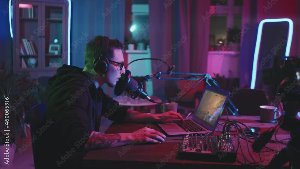 Tracking shot of young man in glasses wearing headphones and talking into microphone while playing video game on laptop in living room lit by neon string lights Tracking shot of young man in glasses wearing headphones and talking into microphone while playing video game on laptop in living room lit by neon string lights