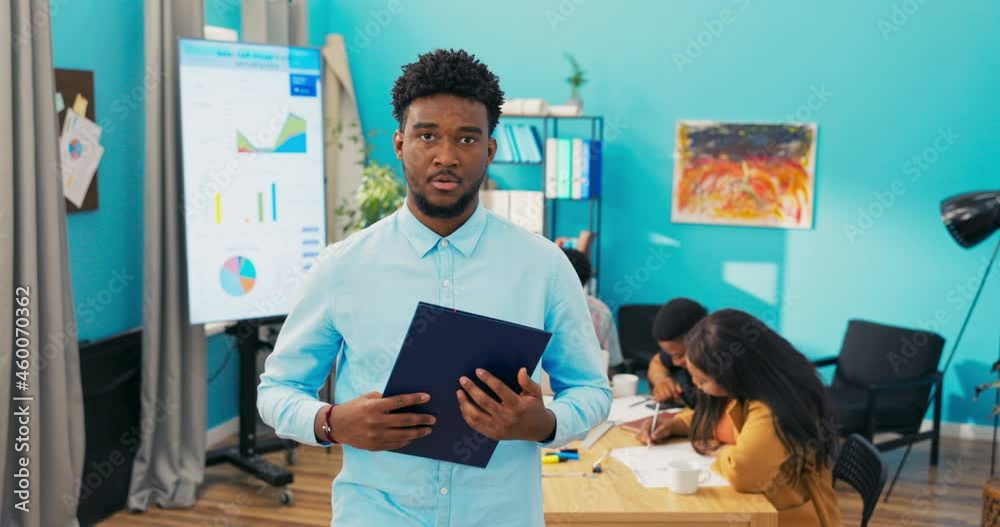 Portrait of a smiling dark-skinned man wearing a shirt with his arms crossed over his chest, the guy has dark hair and stubble, a business meeting of a group of employees is going on in the background
