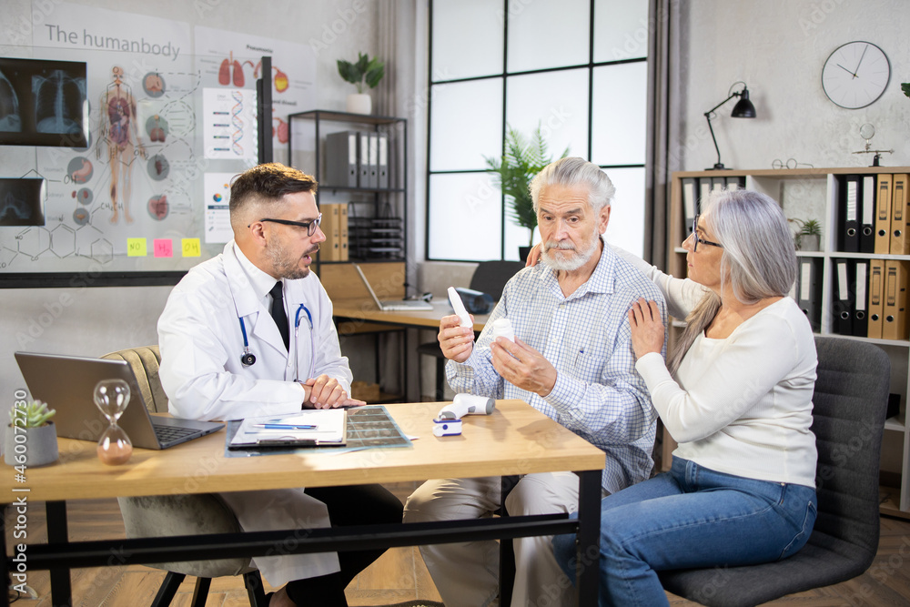 Obraz premium Mature caucasian woman hugging and cheering her sick husband that sitting at doctor's cabinet with various pills in hands. Competent medical worker prescribing treatment for patient.