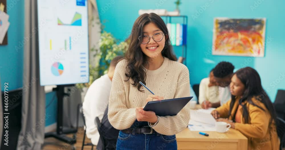 Portrait of Asian woman standing with hands busy with documents, wearing sweater and glasses, smiling. In background view of office, interactive whiteboard, coworkers working on project at table. Portrait of Asian woman standing with hands busy with documents, wearing sweater and glasses, smiling. In background view of office, interactive whiteboard, coworkers working on project at table.