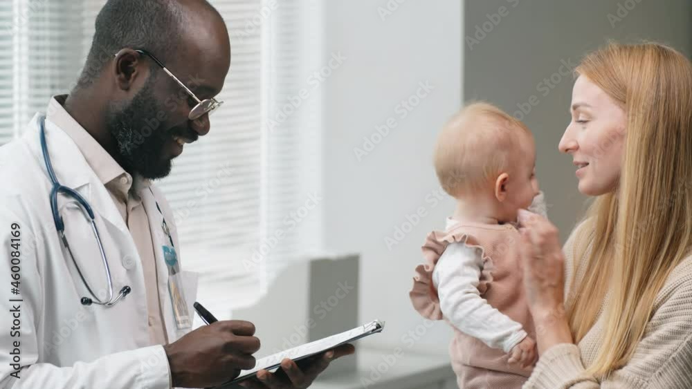 Caucasian woman holding her baby daughter on arms and speaking with African American male doctor as he writing down notes during consultation in clinic Caucasian woman holding her baby daughter on arms and speaking with African American male doctor as he writing down notes during consultation in clinic