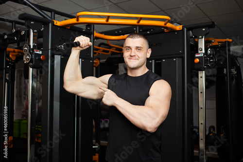 young handsome coach in a black uniform against background of a simulator in gym. Muscular athletic body of a bodybuilder, coaching, individual sports and weight loss Portrait. muscles on the arm