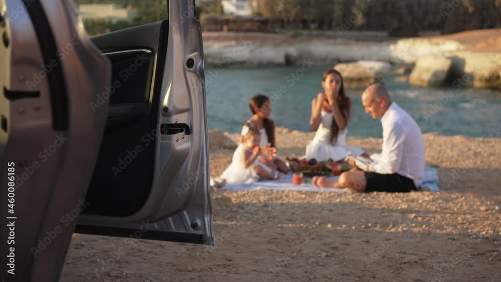 Close-up car with open door and blurred happy Caucasian family talking laughing enjoying family picnic at golden sunset on sandy sea beach. Relaxed confident daughters and father having fun resting Close-up car with open door and blurred happy Caucasian family talking laughing enjoying family picnic at golden sunset on sandy sea beach. Relaxed confident daughters and father having fun resting