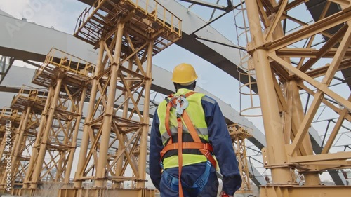 Low angle shot of unrecognizable African-American male worker in hard hat and safety belt with harness walking at construction site with unfinished building with scaffolding photo