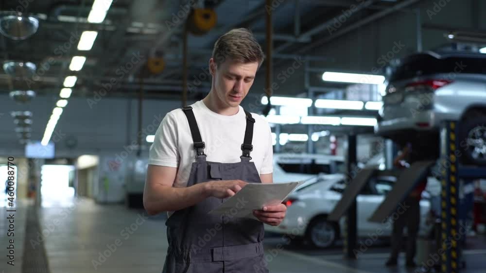 Medium shot portrait of young mechanic male in uniform reading insurance claim form document paper, before maintenance and fixing car for customer in auto repair shop garage with vehicle background. Medium shot portrait of young mechanic male in uniform reading insurance claim form document paper, before maintenance and fixing car for customer in auto repair shop garage with vehicle background.
