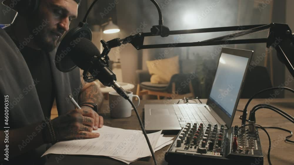 Tracking shot of bearded man sitting at table in dimly lit living room and making notes on his script while talking into microphone and recording podcast