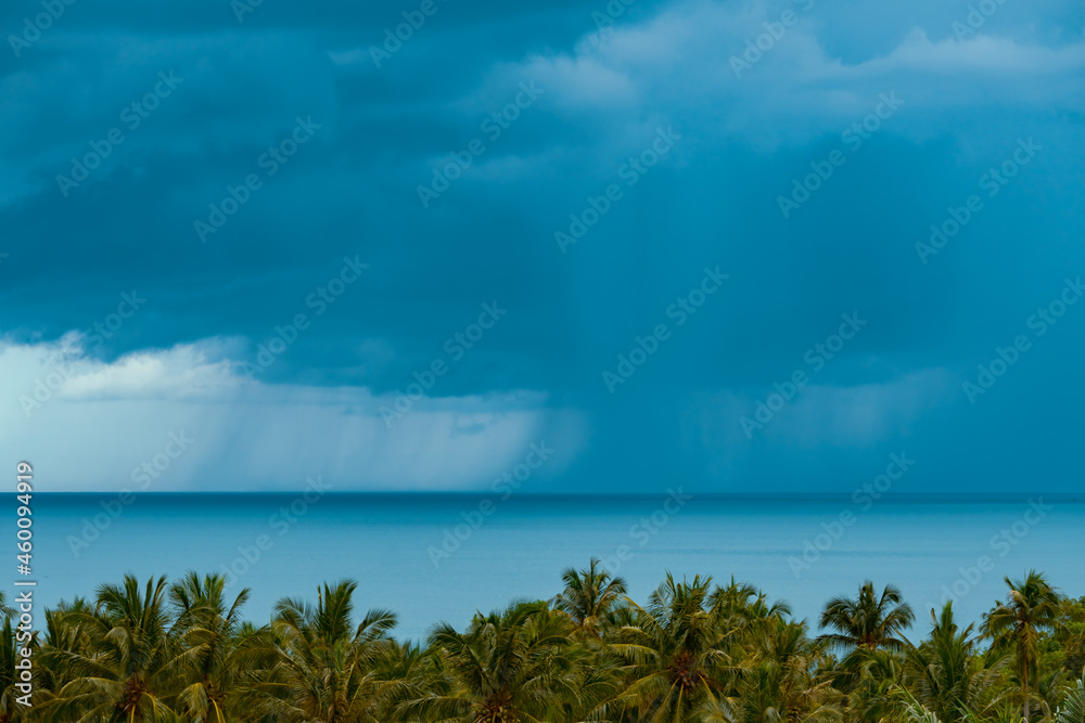 Rain and dramatic clouds over turquoise sea. Palm trees in the foreground.