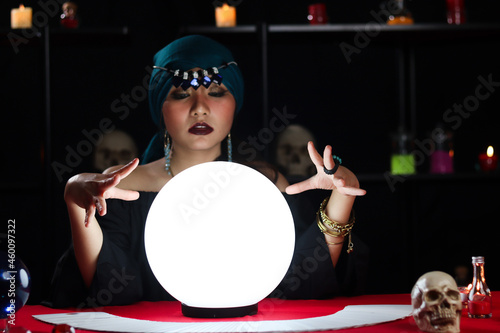 Luminous crystal ball and cards on magnificent fortune telling table with mysterious beautiful woman fortune teller in black dress, dark witch try to read future on magical ball. photo