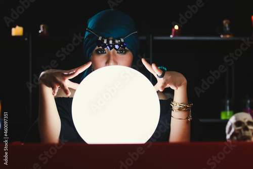 Mysterious magnificent beautiful woman fortune teller in black dress reading future on luminous crystal ball, dark witch casting a spell with magical ball.